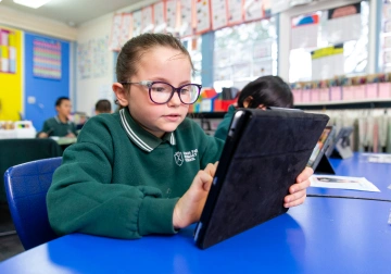 A young girl wearing glasses focused on a tablet at her desk in a classroom