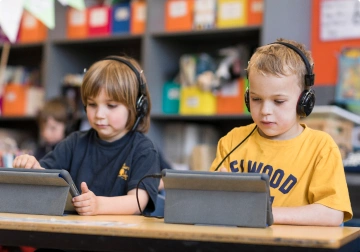 Two young boys wearing headphones working on tablets side by side in a library